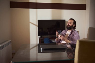 Masa başında sakallı düşünceli bir adam huzurlu bir ortamda içki içiyor. Oda hafifçe aydınlatılmış, sakin ve yansıtıcı bir ortam yaratıyor..