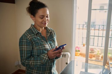 Rahat bir evde oturan bir kadın akıllı telefonunu kontrol eder, sıcak bir şekilde gülümser. Pencereden içeri güneş ışığı giriyor. Huzurlu ve hoş bir atmosferi vurguluyor..