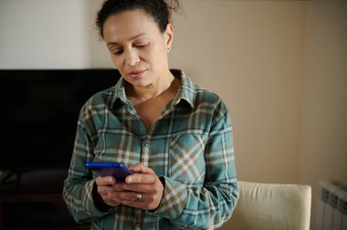 Bir kadın ev ortamında akıllı telefonunu kullanırken görülüyor, düşünceli görünüyor. Gündelik ve rahat kıyafetler giyiyor, modern yaşam tarzını yansıtan ev ortamında konumlandırılmış..