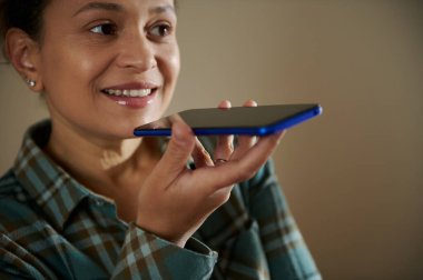 Akıllı telefon tutan bir kadın, ses asistanı özelliğiyle meşgul, içeride, mutlu bir şekilde gülümsüyor..