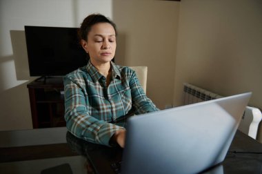 Ekoseli tişört giyen bir kadın ofis masasında dizüstü bilgisayarla çalışıyor..