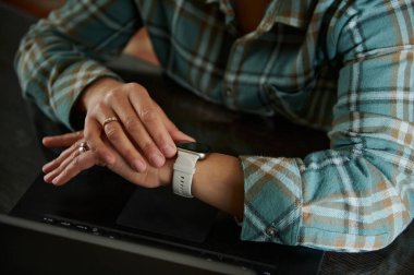 Dizüstü bilgisayarın üzerindeki akıllı bir kasette dinlenen yakın plan bir el. Bireyler rahat bir ofis ortamını vurgulayan ve modern teknoloji kullanımını sembolize eden fanila bir gömlek giyerler..