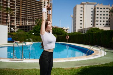 Yeşillik ve modern binalarla çevrili bir havuzun yanında bir kadın kollarını gökyüzüne doğru uzatıyor. Hava güneşli ve açık, pozitif ve sakin bir ortam yaratıyor..