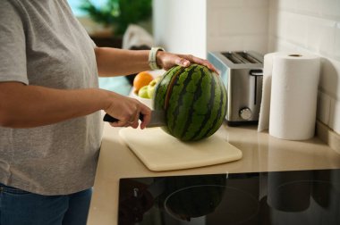 Bir insan mutfak tezgahında olgun bir karpuzu doğruyor, ev eşyaları ve rahat bir atmosferle çevrili, sağlıklı beslenme ve ev hayatını ön plana çıkarıyor..