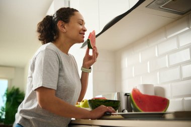 Bir kadın, sağlıklı ve neşeli bir yaşam tarzını yansıtan taze bir karpuz diliminin tadını çıkarır. Ev hayatı ve refah bu parlak mutfak anında kapsülleşir..
