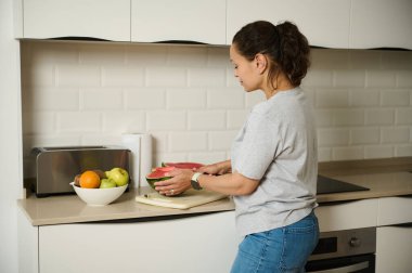 Bir kadın mutfak tezgahında bir kase taze meyveyle meyve dilimliyor, rahat ve modern bir ev ortamında sağlıklı bir yaşam tarzını gösteriyor..