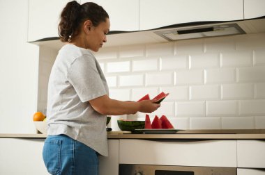 Gündelik giyinmiş bir kadın modern bir mutfakta karpuz dilimleri hazırlıyor. Ev hayatını ve sağlıklı beslenmeyi ön plana çıkarıyor. Ayar gösterileri basitlik ve ev kavramı.