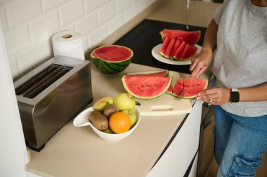 Tezgahta taze meyvelerle çevrili bir kesme tahtasının üzerinde karpuz dilimleyen birinin olduğu bir mutfak sahnesi..