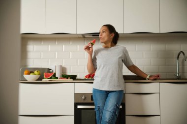 Çağdaş bir mutfakta bir parça taze karpuzun tadını çıkaran bir kadın. Temiz tasarım canlı meyvelerle çelişiyor, sağlıklı bir yaşam tarzı ve huzurlu bir ev hayatı sağlıyor..