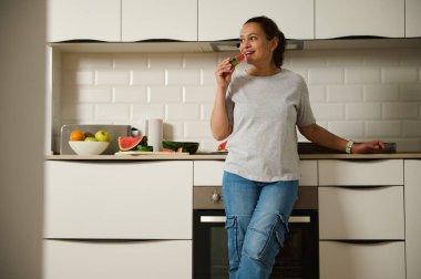 Bir kadın mutfağında durmuş taze meyvelerle çevrili bir dilim karpuz yiyor. Sağlıklı bir yaşam tarzı ve rahat bir ev ortamını resmediyor..