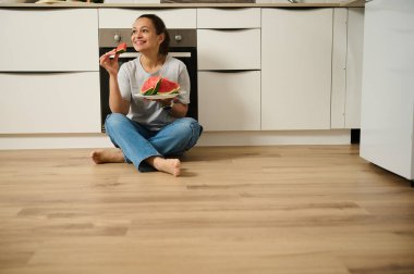 Neşeli bir kadın mutfak zemininde rahatça oturur taze karpuzun tadını çıkarır, gününün tadını çıkarır..