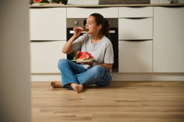 Bir kadın mutfak zemininde oturmuş taze karpuz dilimlerinin tadını çıkarıyor. Parlak iç mekan ve sağlıklı atıştırmalıklar rahat ve rahatlatıcı bir ortam yaratarak basit zevkleri ve ev hayatını ön plana çıkarıyor..