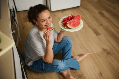 Bir kadın mutfak zemininde oturmuş taze karpuz dilimleri yerken gülümsüyor. Bu an bir neşe, rahatlama ve evcil bir ortamda sağlıklı bir yaşam hissi yakalıyor..