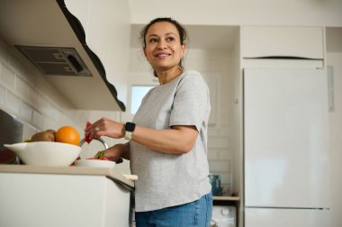 Neşeli bir kadın taze ürünlerle çevrili mutfağında yemek hazırlıyor. Parlak mekan ve iyimser tavrı sağlıklı bir yaşam ve aile odaklı bir aile hayatı hissi veriyor..