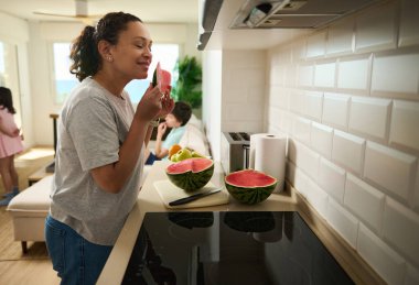 Bir kadın mutfakta, ailesi evde birlikte vakit geçirirken taze bir karpuzun tadını çıkarıyor. Ortam, aile ve sağlıklı beslenmeye odaklanmış sıcak ve evcil bir ortamı yansıtıyor..
