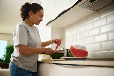 Bir kadın mutfak tezgahında karpuz dilimliyor, ev hayatını ve sağlıklı alışkanlıklarını gösteriyor. Parlak ve temiz iç mekan, konforlu ve pratik bir aile yaşam tarzını vurguluyor.