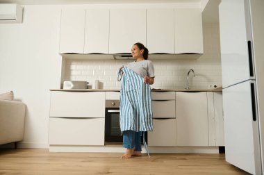 Bir kadın parlak, modern bir mutfakta elinde çizgili bir önlükle duruyor. Yerleşim, ev hayatı, konfor ve aşçılığa hazırlık hissi veriyor ve ev ortamını vurguluyor..