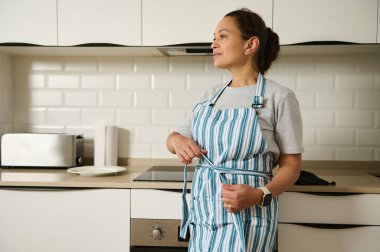 Çizgili önlük giyen bir kadın modern bir mutfakta rahatça durur. Önlüğü bağlıyor, yemek pişirmeye ya da ev işi yapmaya hazırlandığını gösteriyor..