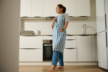 Neşeli bir kadın modern bir mutfakta çıplak ayakla duruyor, önlük ve günlük giysiler giyiyor. Sahne ev yemeklerinin özünü yakalıyor, rahatlatıcı ve davetkar bir atmosfer yaratıyor..