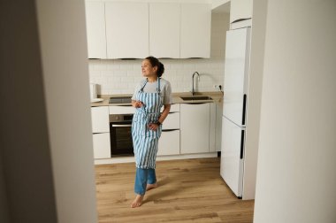 Bir kadın modern mutfağında duruyor, gülümsüyor ve neşeli görünüyor. Mutfak parlak ve minimalist. Önlük takıyor, yemek pişirmeyi veya yaratıcılığı öneriyor..