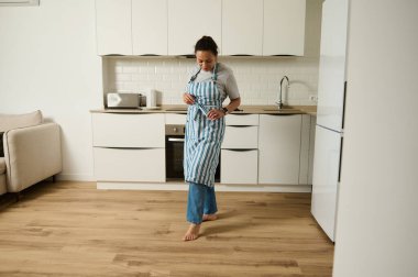 Şık çizgili önlüklü bir kadın yemek pişirmeye hazır minimalist bir mutfakta duruyor. Modern ev dekorasyonu sakin, evcil bir his yaratır..