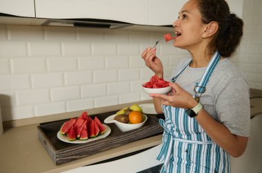 Çizgili önlüklü bir kadın modern, parlak bir mutfakta dururken karpuz yer. Sahne, ev ortamında sağlıklı yaşam, tazelik ve konforun özünü yansıtıyor..