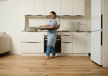 Önlük takan mutlu bir kadın, mutfak ortamında yemek tepsisi taşır. Parlak mekan ve gülüşü, ev hayatını ve ev yapımı mutfağı ön plana çıkaran sıcaklık ve neşeyi çağrıştırıyor..