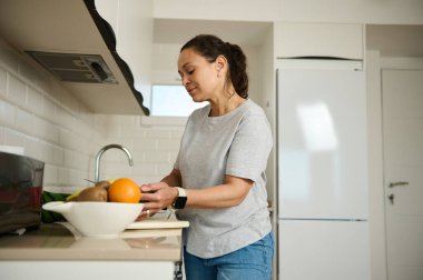 Bir kadın, modern bir mutfakta sağlıklı bir yemek hazırlarken taze meyve seçiyor. Parlak, düzenli iç ve doğal gıda malzemeleri sağlıklı bir yaşam ve farkındalık hissi uyandırıyor..
