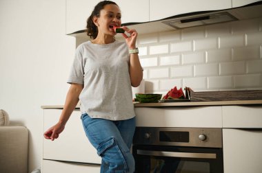 Neşeli bir kadın mutfak tezgahına yaslanır ve bir dilim karpuzun tadını çıkarır. Modern çevre, taze meyve ve onun rahat duruşu sıcak, yaşam tarzı odaklı bir an yaratır..