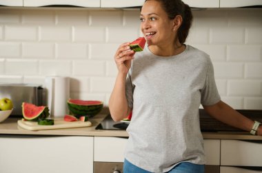 Neşeli bir kadın parlak bir mutfakta durup bir dilim taze karpuzun tadını çıkarıyor. Atmosfer, modern bir ev ortamında sıcaklık, sağlık ve rahatlama duygusu taşır..