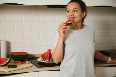 Bir kadın sıcak bir mutfakta ferahlatıcı bir karpuzun tadını çıkarırken çekilen neşeli bir an. Ayar, sağlıklı ve rahat bir yaşam tarzı sergiliyor. Taze meyveler refahı vurguluyor..