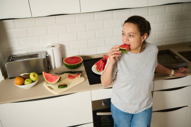 Bir kadın mutfağında durup bir dilim taze karpuz yiyor. Sahne, evde taze meyvelerin keyfine dikkat çekerek ev içi rahatlık ve sağlıklı bir yaşam sağlıyor..