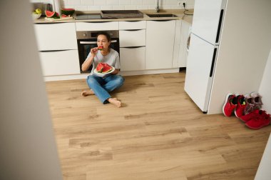 Genç bir kadın modern bir mutfağın ahşap zemininde oturmuş tabaktan taze karpuz dilimlerinin tadını çıkarıyor. Sahne gündelik yaşama, sağlıklı yeme ve ev içi rahatlık hissi veriyor..