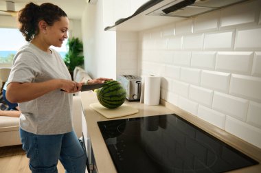 Neşeli bir kadın evde karpuz hazırlıyor, sıcak ve canlı bir ev sahnesi sergiliyor. Parlak mutfak ve olumlu tavrı sağlıklı yaşam ve aile rahatlığını çağrıştırıyor..