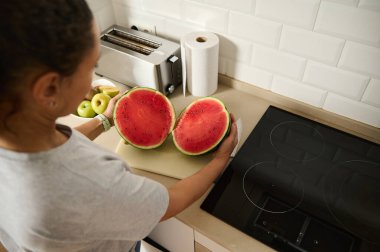 Bir kadın, parlak ve modern bir mutfak ortamında taze kesilmiş bir karpuzun iki yarısını tutuyor. Sahnede ev hayatı, taze yemek hazırlama ve sağlıklı bir yaşam tarzı sergileniyor..