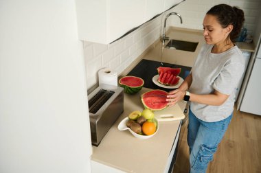 Parlak bir mutfakta taze meyvelerle karpuz doğrayan bir kadın. Sahne, rahat ve modern bir ortamda sağlıklı yemek pişirme, ev hayatı ve taze yemek hazırlama görüntülerini yakalıyor..