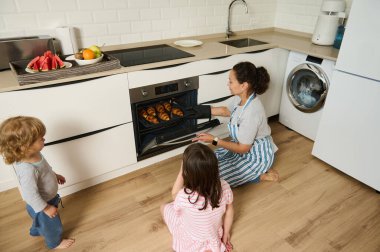 Bir anne, çocukları bakarken modern bir mutfakta kruvasan pişirir. Parlak bir ev ortamında sağlıklı yemek ve kahvaltı hazırlıklarında sıcak bir aile ortamı yaratır..