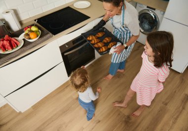Bir anne, modern bir mutfakta çocuklarına taze kruvasan servis eder. Atmosfer neşeli ve sıcak. Yemek hazırlıkları sırasında aile bağlarını yansıtıyor..