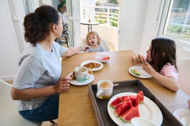 Bir anne ve iki çocuk, aydınlık ve modern bir mutfakta sağlıklı bir kahvaltıyı paylaşıyorlar. Temiz tasarım ve manzaralı balkon, taze meyve ve kruvasanların servis edildiği sakin sabah atmosferini vurguluyor..