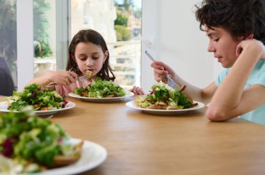 Bir erkek ve kız kardeş, modern ve parlak bir mutfakta taze salata yer. Güneşli gün, aile atmosferine sıcaklık katar. Yemek sağlıklı beslenmeye ve refaha odaklanır..