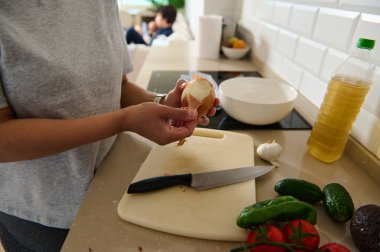 Çağdaş bir mutfakta domates ve salatalık da dahil taze malzemelerle soğan soyan bir insan. Mutfak hazırlığını ve sağlıklı beslenmeyi vurgulayan detaylı bir bakış açısı.