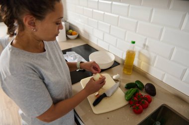 Modern bir mutfakta kesme tahtasının üzerinde taze sebzeler hazırlarken bir kadın soğan soyuyor. Domates, avokado, salatalık, sarımsak ve zeytinyağı gibi malzemeler düzgünce düzenlenmiştir..