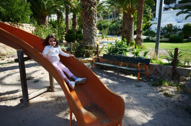 Gülümseyen bir kız canlı bir parkta turuncu bir kaydırağın keyfini çıkarıyor. Sahne neşeyi, yeşilliği ve güneşli bir günü bu canlı açık hava oyun bahçesinde çocukluk eğlencesi ve doğayı yansıtıyor..