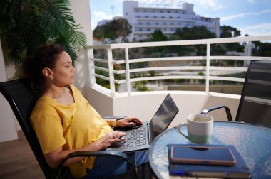 Bir kadın balkonda otururken işe odaklanıyor, dizüstü bilgisayarı kucağında ve yakınlardaki bir masada kahve fincanı ile güneşli bir açık hava ortamının tadını çıkarıyor. Huzurlu ortama tropikal bir hava eşlik ediyor..