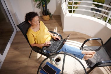 Bir kadın, güneşli ve huzurlu bir günü kucaklayarak, günlük kıyafetleriyle balkonda uzanırken uzaktan bilgisayarla çalışmaktan hoşlanır..