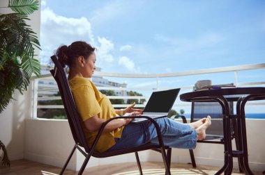 Bir kadın, okyanus manzaralı bir balkonda dizüstü bilgisayar üzerinde çalışırken rahat bir şekilde rahatlar, sakin, sakin ve ilham verici bir çalışma ortamında üretkenliği somutlaştırır..