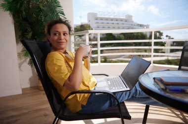 Bir kadın, dizüstü bilgisayarla çalışırken ve güneşli bir balkonda kahve içerken boş zamanlarından zevk alır. Rahatlama, uzak çalışma ve açık hava yaşam tarzı temaları için ideal.