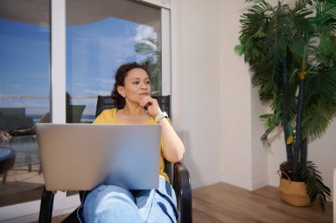 Evde oturan bir kadın, rahat bir ortamda dizüstü bilgisayarı üzerinde çalışıyor, doğal ışık ve bitkiler huzurlu atmosferi geliştiriyor..