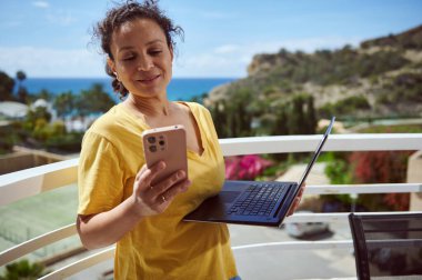 Balkonda elinde akıllı telefon ve dizüstü bilgisayarla deniz manzarası ve yemyeşil manzaranın tadını çıkaran bir kadın. Modern teknolojiyi ve barışçıl uzaktan çalışmayı simgeliyor..