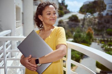 Mutlu bir kadın terasta duruyor, dizüstü bilgisayarı kucaklıyor, günlük giysiler giyiyor ve dışarıda eğleniyor. Güneşli bir gün ve rahat bir atmosfer memnuniyet ve pozitifliği iletir..